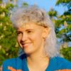 Eine junge Frau mit lockigem, wei&szlig;blondem Vokuhila-Haarschnitt, die an einem sonnigen Tag drau&szlig;en sitzt.