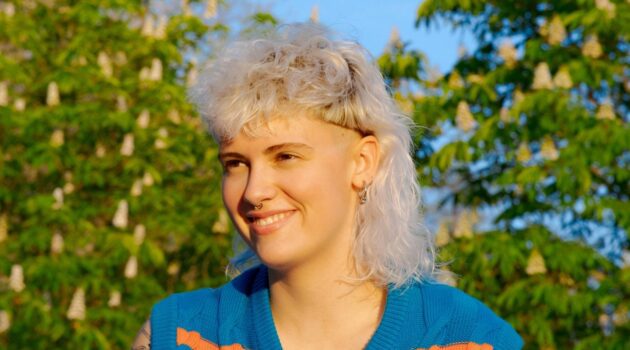Eine junge Frau mit lockigem, weißblondem Vokuhila-Haarschnitt, die an einem sonnigen Tag draußen sitzt.