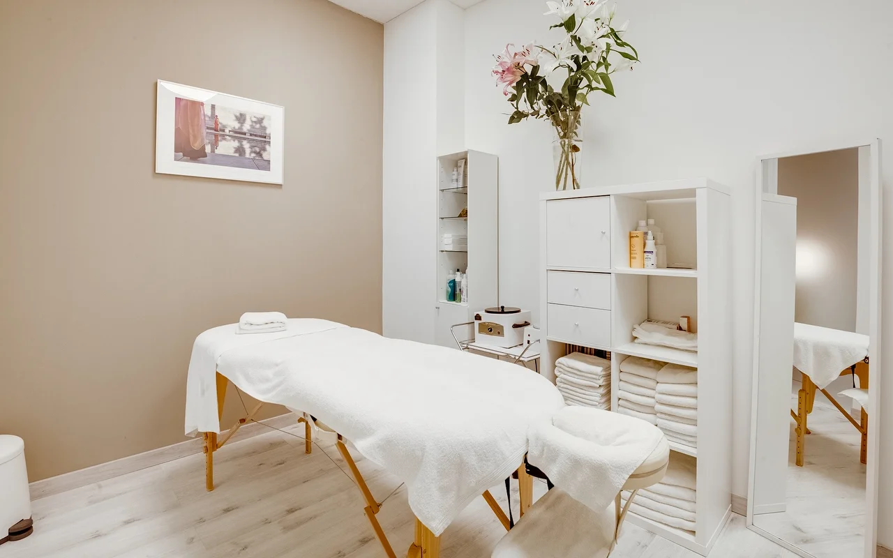 Ein ruhiger Behandlungsraum mit einer weißen Massageliege und frischen Handtüchern. Der Raum hat neutrale Beige- und Weißtöne, einen weißen Schrank mit gefalteten Handtüchern und eine hohe Glasvase mit weißen und rosafarbenen Lilien.