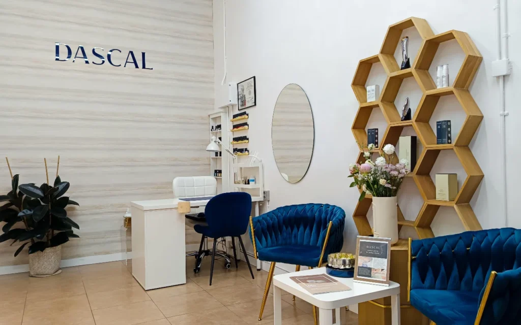 Ein stilvolles Beauty-Salon-Interieur mit tiefblauen Samtsesseln und einem sechseckigen Holzregal an der Wand. Ein weißer Maniküretisch steht in der Ecke an einer hellen, texturierten Wand mit dem Schriftzug „DASCAL“ in blauen Buchstaben.