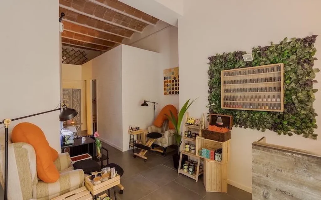 Ein gemütliches Nagelstudio im Bohème-Stil mit weichen Sesseln und orangefarbenen Kissen. An der Wand eine große vertikale Pflanzeninstallation rund um ein Holzregal voller Nagellackflaschen.