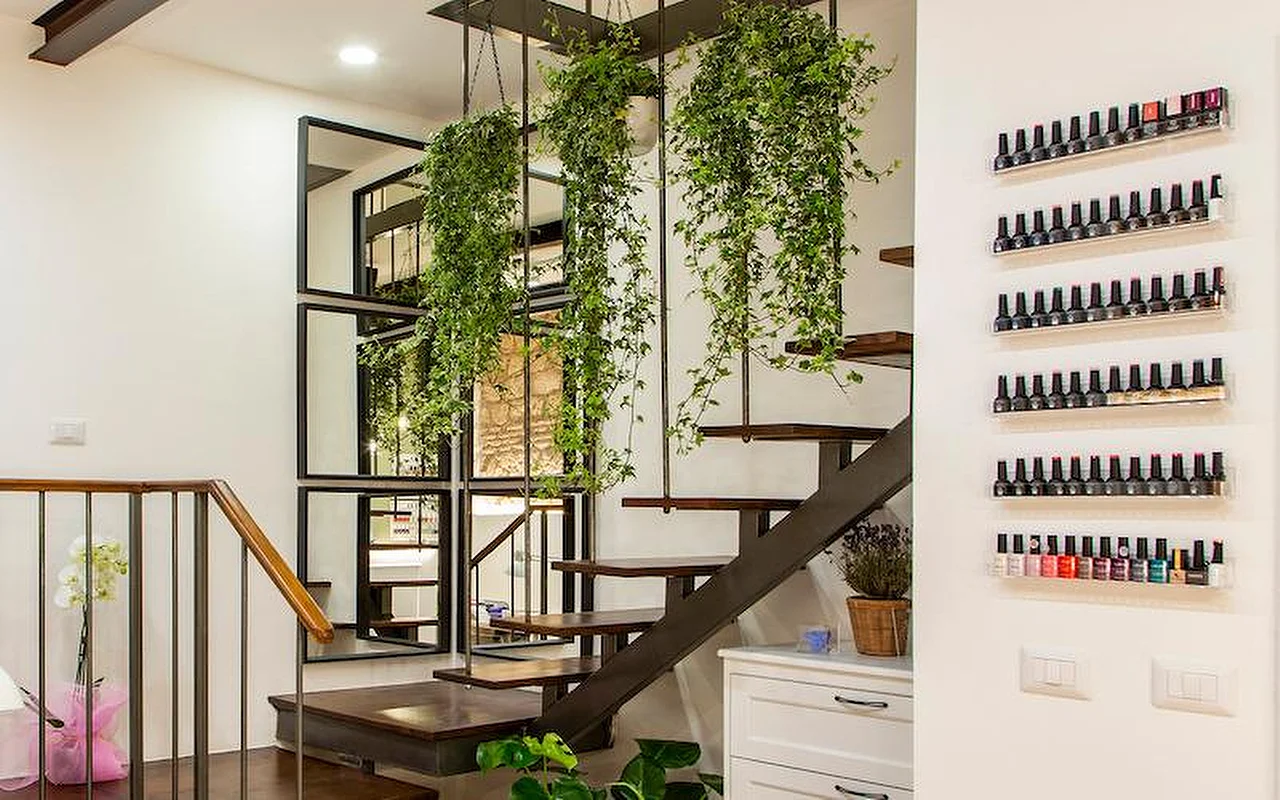 Ein stilvolles, modernes Interieur mit einer freitragenden Treppe aus Holz und Metall. Üppige Hängepflanzen ranken von der Decke zwischen den Stufen, und Reihen bunter Nagellackflaschen stehen auf schlanken, transparenten Regalen an der weißen Wand.