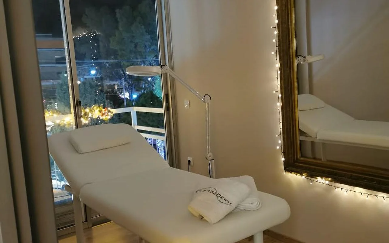 Ein sanft beleuchteter Behandlungsraum mit einem weißen Bett, einem CasaDerm-Handtuch, einer Vergrößerungslampe, Lichterketten um einen goldenen Spiegel und einem Balkonfenster mit Blick auf die Glyfada-Straßenszene am Abend.