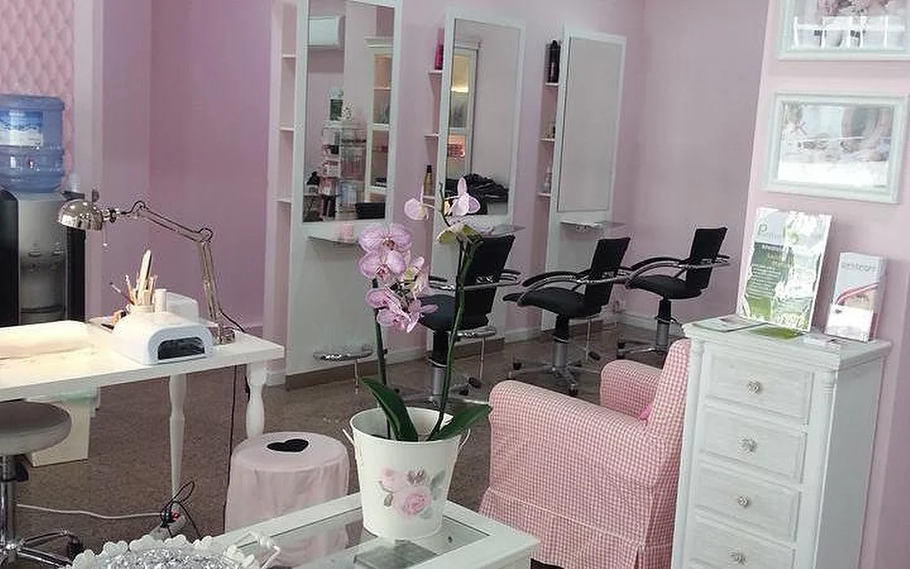 Das Innere eines hellen, in Pink gehaltenen Haar- und Nagelsalons. Links eine weiße Manikürestation mit Schreibtischlampe und UV-Nagelgerät. In der Mitte eine Orchidee in einem weißen Topf auf einem Couchtisch neben einem rosa-weiß karierten Sessel. Im Hintergrund mehrere Frisierstationen mit hohen Spiegeln und schwarzen Salonsesseln vor hellrosa Wänden.