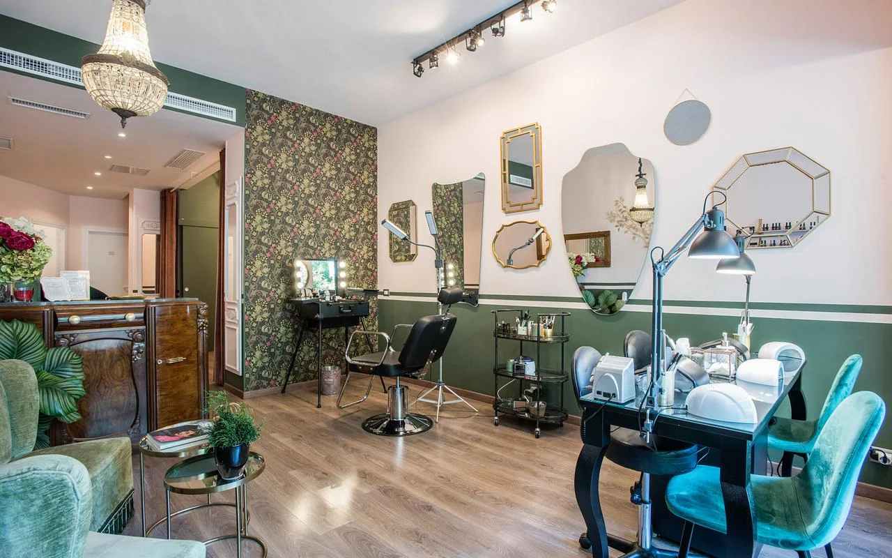 Ein Salon im Vintage-Chic-Stil mit weißen und waldgrünen Wänden. Blumentapete, eine Sammlung unregelmäßig geformter, goldgerahmter Spiegel, eine professionelle Make-up-Station und üppige grüne Samtsitze.