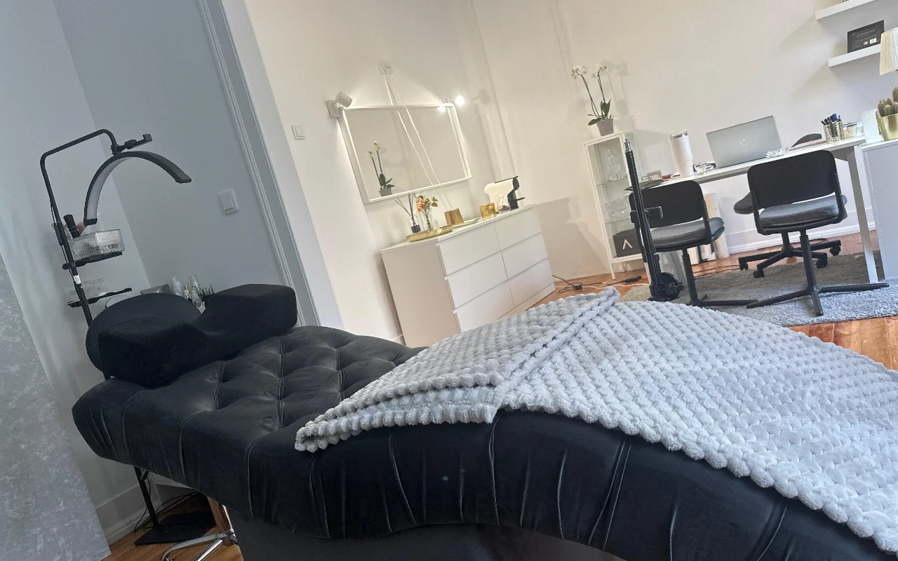 Ein moderner Raum für Wimpernbehandlungen aus niedriger Perspektive. Im Vordergrund eine gepolsterte schwarze Behandlungsliege mit einer strukturierten grauen Decke und einem ergonomischen Nackenstützkissen unter einer gebogenen Ringleuchte. Im Hintergrund ein minimalistischer weißer Arbeitsplatz mit Schreibtisch, zwei schwarzen Stühlen, Laptop, weißer Kommode und großem Spiegel.