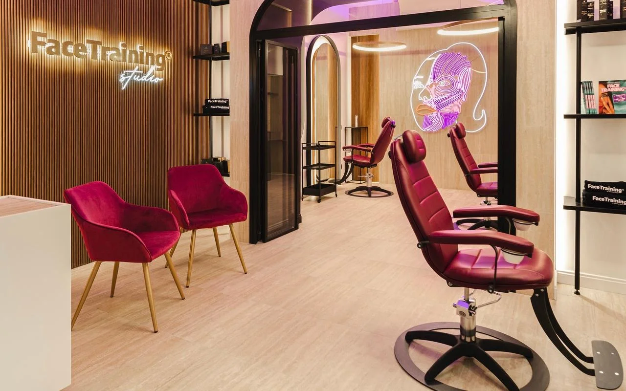 Ein minimalistisches Studio mit einer Holzlatten-Akzentwand und hintergrundbeleuchtetem Logo. Der Behandlungsbereich umfasst professionelle burgunderrote Lederstühle und eine Neonlicht-Installation, die ein stilisiertes Gesichtsmuskeldiagramm auf einer Glastrennwand zeigt.