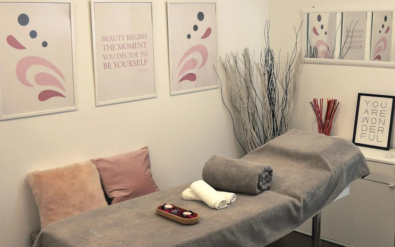 Ein heller Behandlungsraum mit einer Massageliege, die mit einem grauen Handtuch bedeckt ist. Die Wand ist mit abstrakter Kunst in Pastelltönen und motivierenden Sprüchen dekoriert, während dekorative weiße Zweige und Kerzen eine ruhige Atmosphäre schaffen.