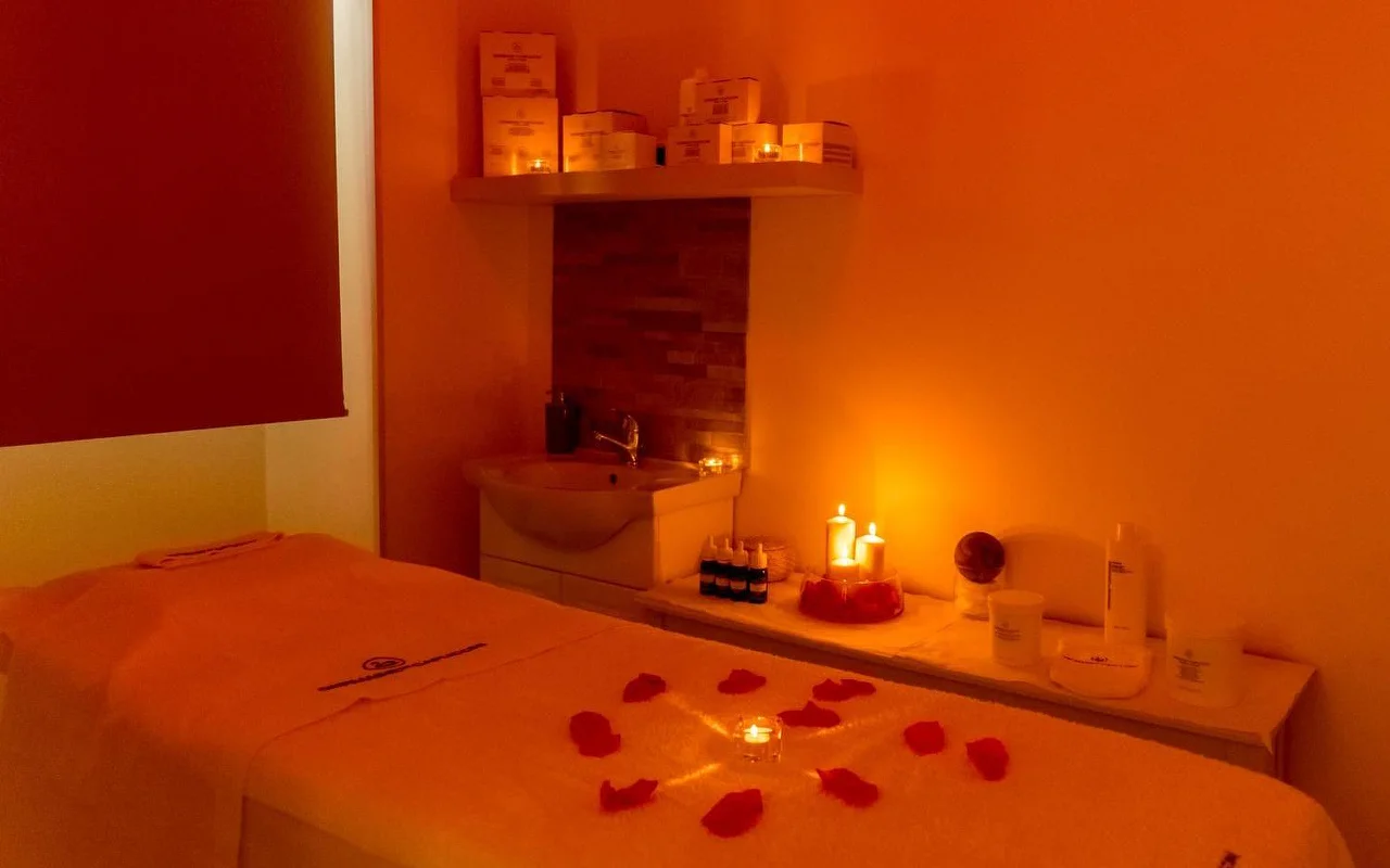 Ein Massageraum mit warmem, stimmungsvollem orangefarbenem Licht und flackernden Kerzen. Rote Rosenblätter sind kreisförmig um eine zentrale Kerze auf der Behandlungsliege arrangiert, mit ordentlich aufgereihten Beauty-Produkten auf einem Regal dahinter.