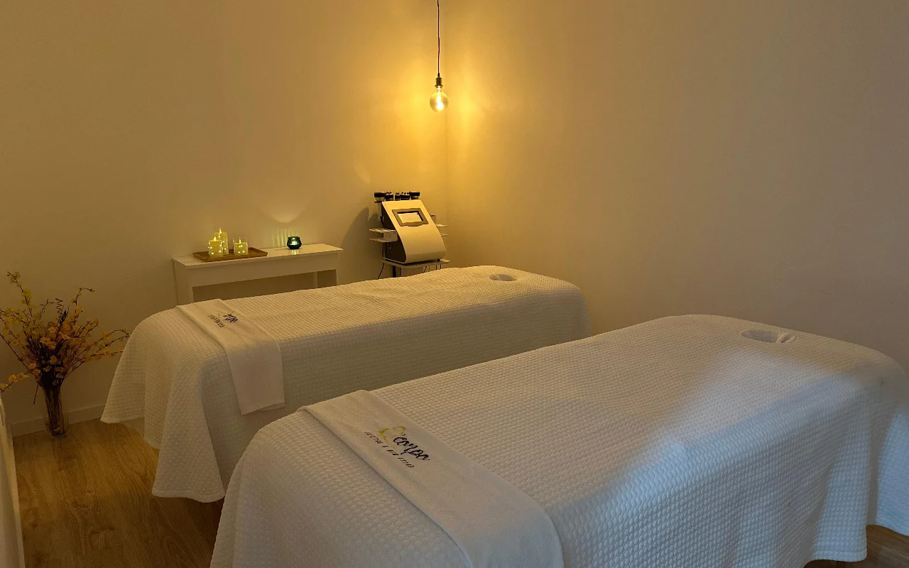 Ein ruhiger Massage- und Behandlungsraum mit zwei nebeneinanderstehenden Behandlungsliegen, bedeckt mit frischen weißen Handtüchern und Laken. Der Raum wird von einer einzelnen warmen Glühbirne und kleinen Kerzen auf einem Beistelltisch beleuchtet, die eine gelassene Atmosphäre schaffen. Im Hintergrund steht ein spezialisiertes Kosmetikgerät zwischen den Liegen.