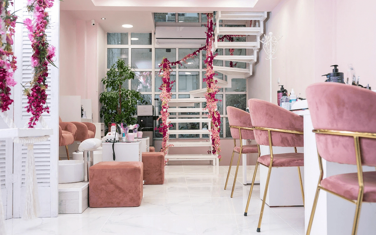 Ein feminines Nagelstudio mit zartrosa Samtsesseln, einer weißen Wendeltreppe mit rosa Kunstblumen, Lamellentrennwänden und goldbeinigen Manikürestationen auf marmorähnlichem Boden.