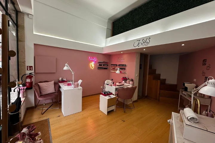 Ein stilvolles Nagelstudio mit hellrosa Wänden und warmen Holzböden. Der Raum enthält mehrere weiße Maniküretische mit altrosa Samtsesseln. Ein leuchtend pinkes Neonschild in Form einer Hand mit Nagellackflasche und dem Schriftzug „Nails“ leuchtet an der Wand. Auf einem Zwischengeschoss ist eine grüne Kunsthecke sichtbar.