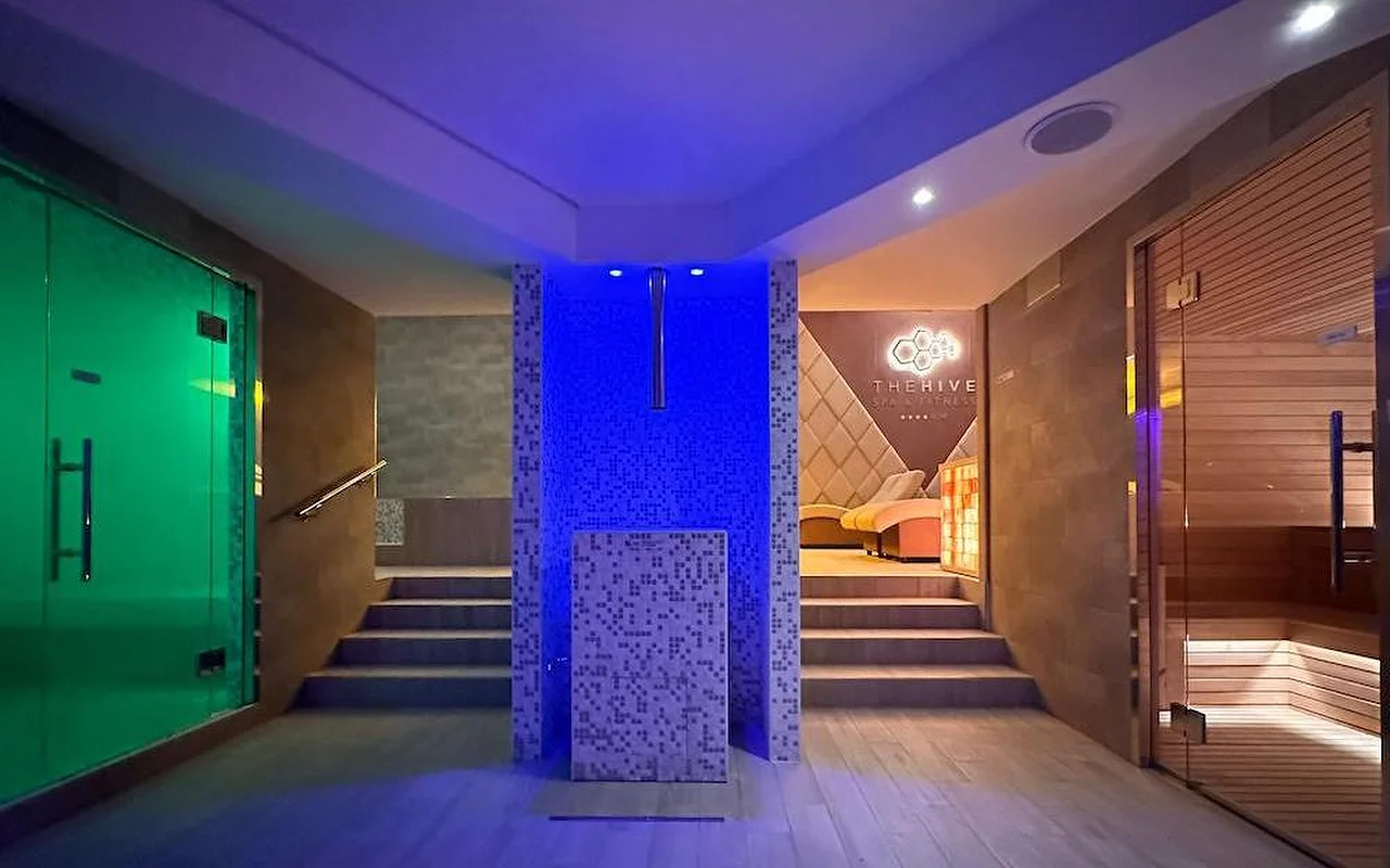 Der Thermalbereich eines Luxus-Spas mit stimmungsvoller blau-grüner Beleuchtung. Der Raum umfasst eine finnische Holzsauna, einen Dampfbad-Eingang und ein zentrales Mosaik-Element mit Stufen, die zu Ruhezonen führen.