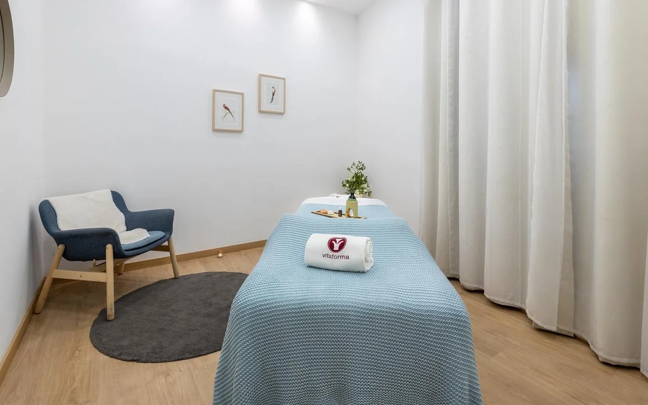 Ein sauberer, minimalistischer Spa-Raum mit weißen Wänden und hellem Holzboden. Eine Behandlungsliege ist ordentlich mit einer hellblauen Strickdecke und einem gerollten weißen Handtuch mit dem Logo „Vitaforma“ hergerichtet. In der Ecke steht ein stilvoller blauer Sessel auf einem kleinen runden grauen Teppich, und lange weiße Vorhänge hängen am Fenster.