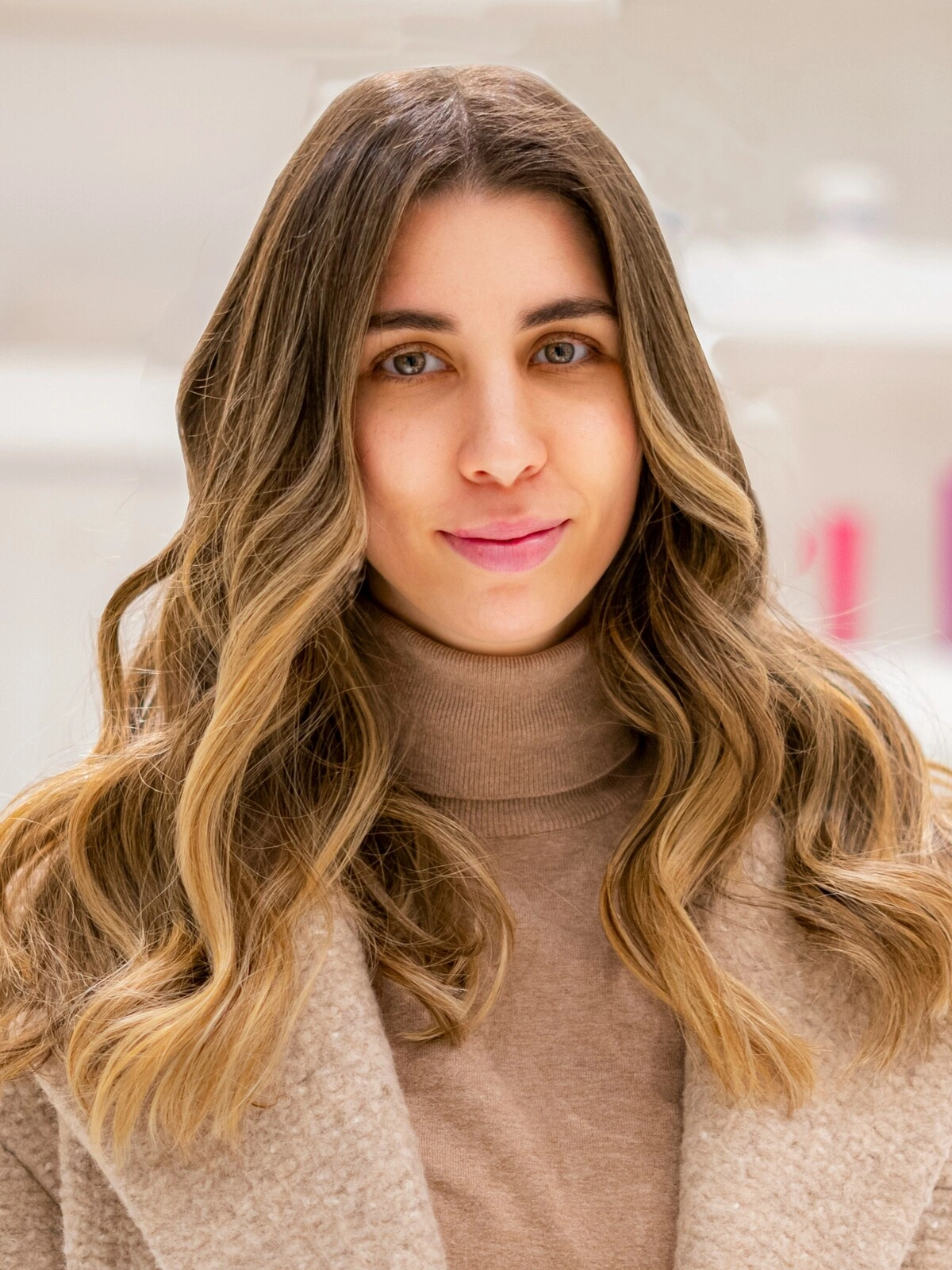 Eine Frau in einem kamelfarbenen Rollkragen und Wollmantel mit schwungvoller, welliger Balayage, bei der brünettes Haar in Karamelltöne übergeht