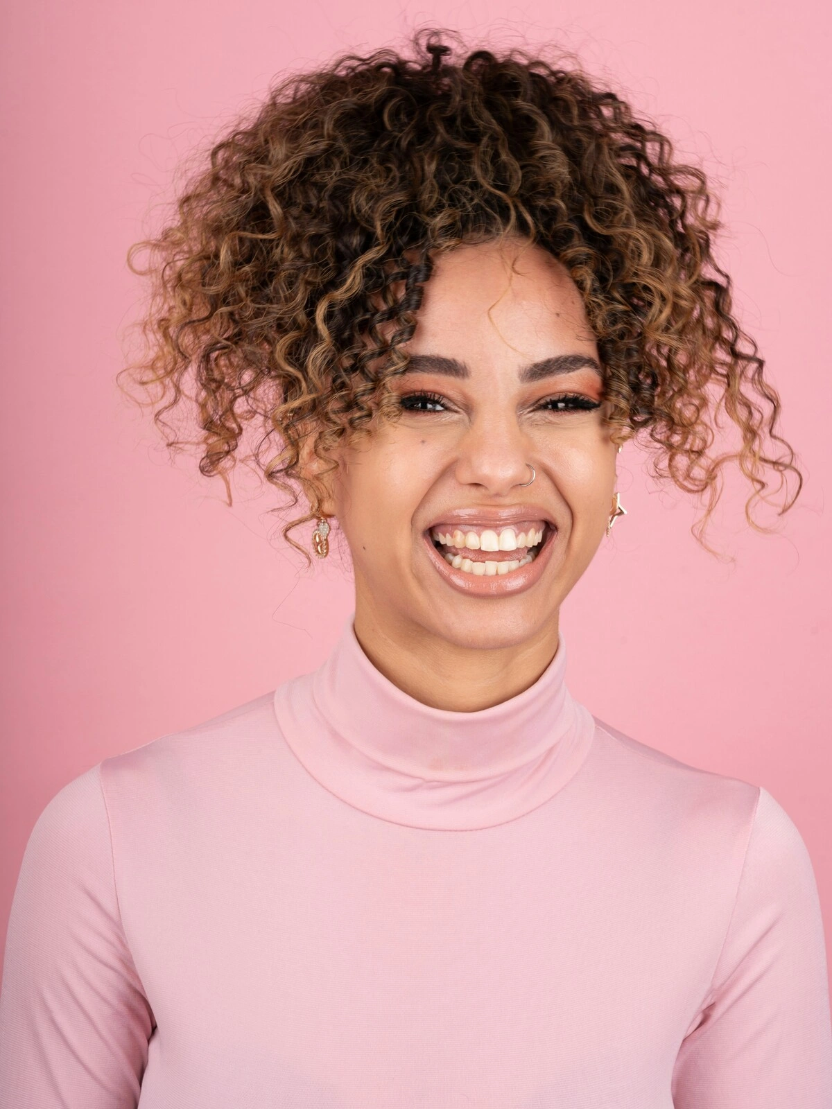 Studioaufnahme einer lächelnden Frau mit Afro-Balayage in einem zartrosa Rollkragenpulli