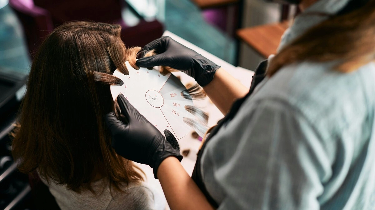 Nahaufnahme einer Coloristin, die Haarsträhnen-Muster an den Kopf einer Kundin hält, um Farbtöne für eine Balayage auszuwählen