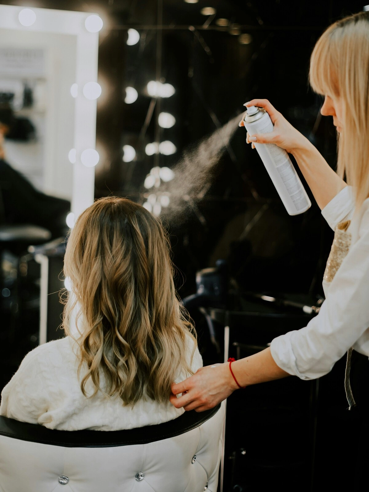 Eine Friseurin sprüht nach einer Balayage, einem Föhnen und dem Styling zu Wellen ein Finish-Produkt ins Haar einer Kundin
