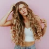Studioaufnahme einer Frau mit sehr langem, welligem, sonnengeküsstem Balayage-Haar, die eine hellblaue Jeans und ein weißes Strick-Crop-Top trägt