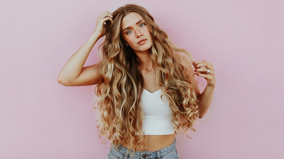 Studioaufnahme einer Frau mit sehr langem, welligem, sonnengeküsstem Balayage-Haar, die eine hellblaue Jeans und ein weißes Strick-Crop-Top trägt