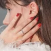 Nahaufnahme der Seitenansicht einer brünetten Frau, die mit der Hand ihr Gesicht berührt und eine glänzende rote Gel-Maniküre zeigt