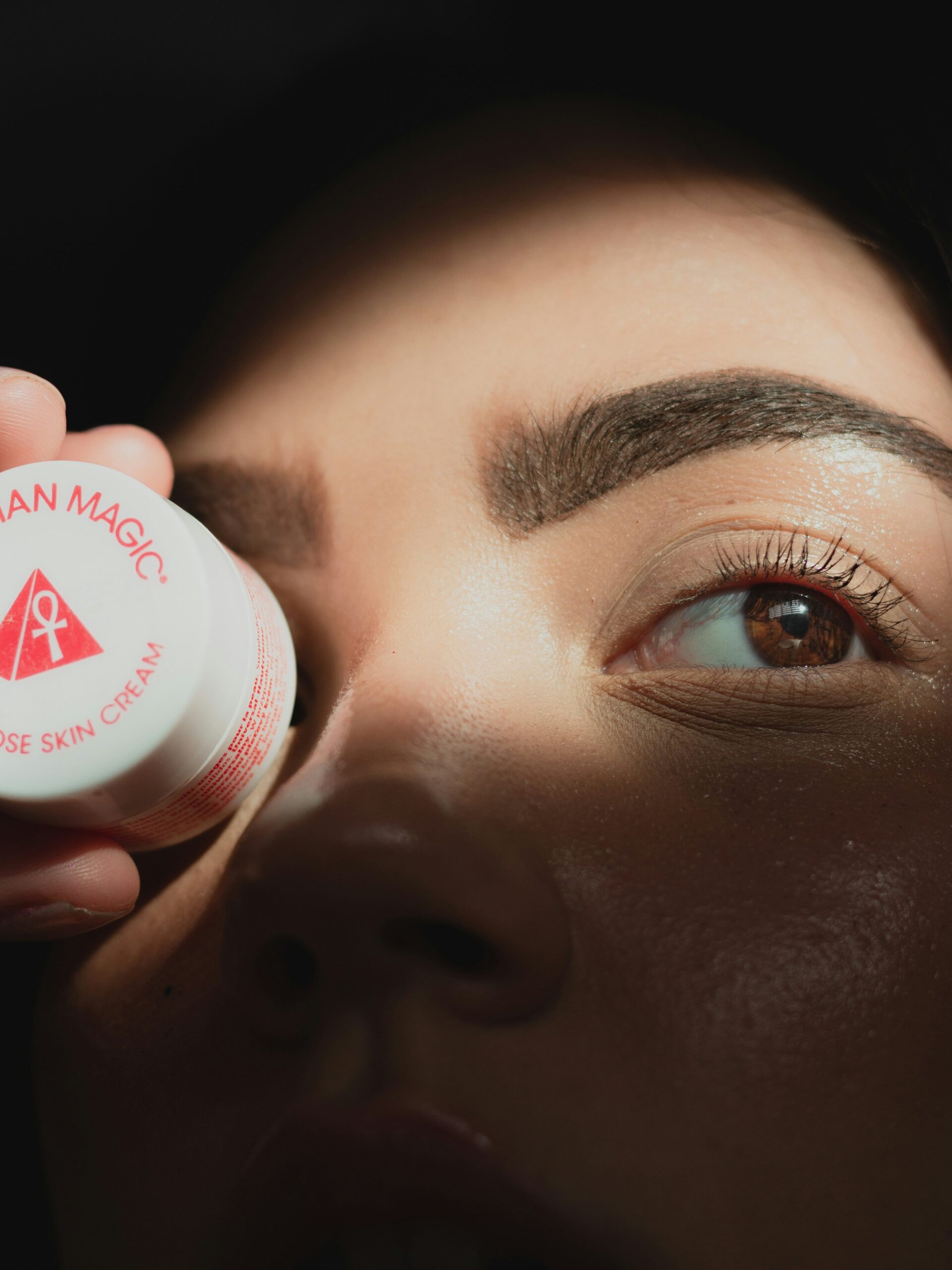 Nahaufnahme des Gesichts einer Frau mit Microblading an den Brauen, die einen kleinen runden Cremetiegel vor ein Auge hält