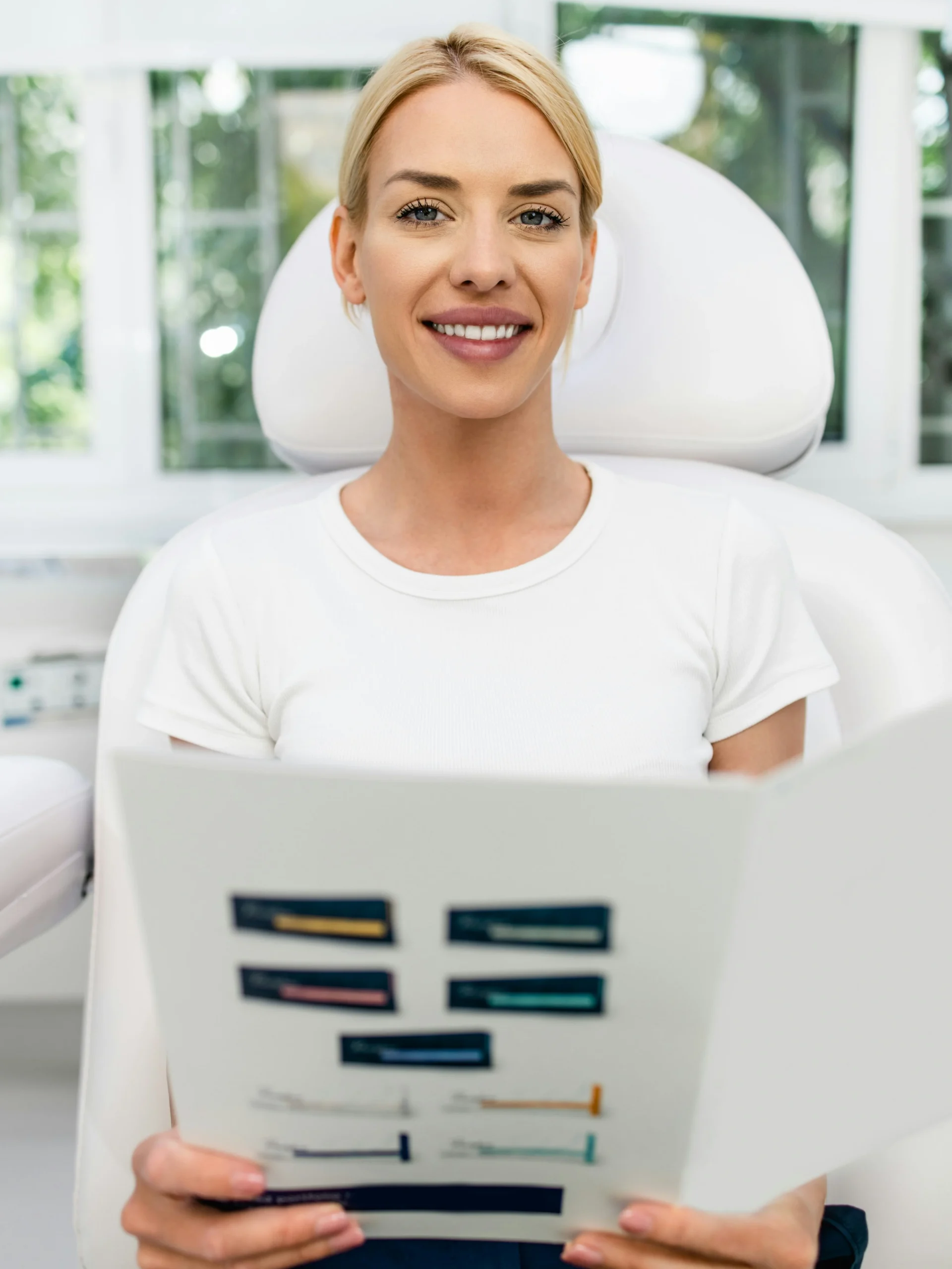 Eine Kundin sitzt in einer hellen, sauberen Klinik und lächelt, während sie eine Informationsbroschüre über Gesichtsbehandlungen in der Hand hält