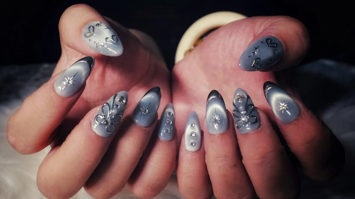 Nahaufnahme der Hände einer Frau mit aufwendigen Acrylnägeln im grauen Cat-Eye-Design, silbernen Herzen und Sternen sowie funkelnden Schmucksteinen