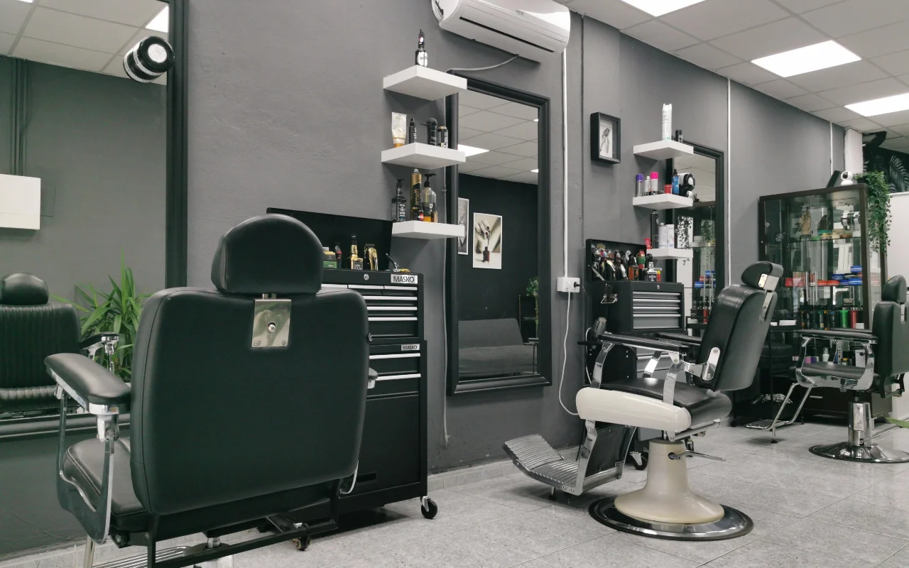 Ein traditioneller Barbershop mit anthrazitgrauen Wänden, klassischen schwarzen Lederbarberstühlen mit Chromfuß und Fußstützen, weißen schwebenden Regalen voller Pflegeprodukte und großen Spiegeln auf gefliestem Boden.
