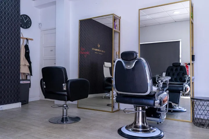 Ein reduziert eingerichteter Salon mit einem schwarzen Stylingstuhl und einem Barberstuhl aus Chrom und Leder auf hellem Holzboden, einem großen goldgerahmten Spiegel, einer Wanduhr und Umhängen, die neben einer dunklen Akzentwand hängen.