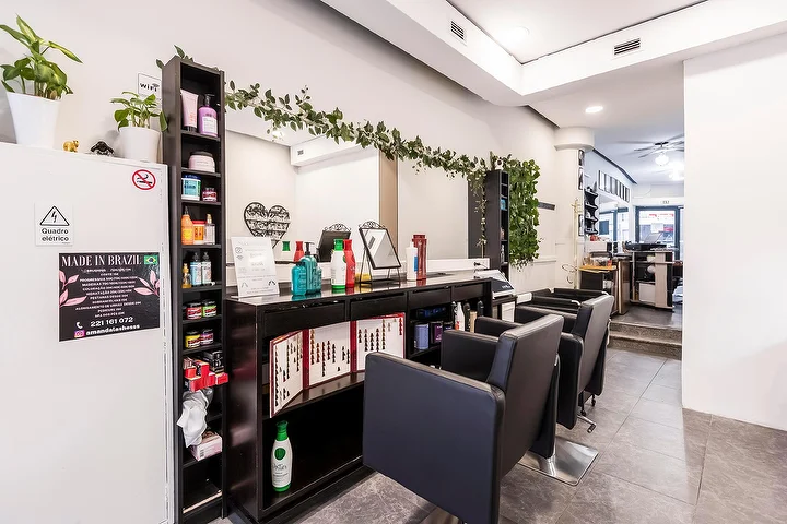 Ein schmaler Friseursalon mit schwarzen Stylingstühlen entlang einer verspiegelten Wand, einem dunklen Regal voller Haarpflegeprodukte, einer herabhängenden Grüngirlande über dem Spiegel und einem gerahmten Made-In-Brazil-Schild auf einer weißen Säule.