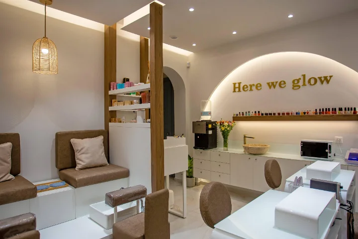 Ein warmer, sanft beleuchteter Nagelsalon mit einer geflochtenen Rattan-Hängelampe, gepolsterten braunen Pediküre-Bänken, weißen Maniküre-Tischen, einem Regal voller bunter Lacke und einem goldenen „Here we glow“-Schriftzug auf einem hinterleuchteten Bogen.