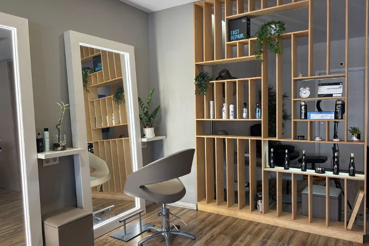 Eine Frisierstation mit grauem Stylingstuhl vor einem gerahmten, LED-beleuchteten Spiegel, einer Regaltrennwand aus Holzlatten mit Stylingprodukten, einer Uhr und Pflanzen, auf Boden in Holzoptik.