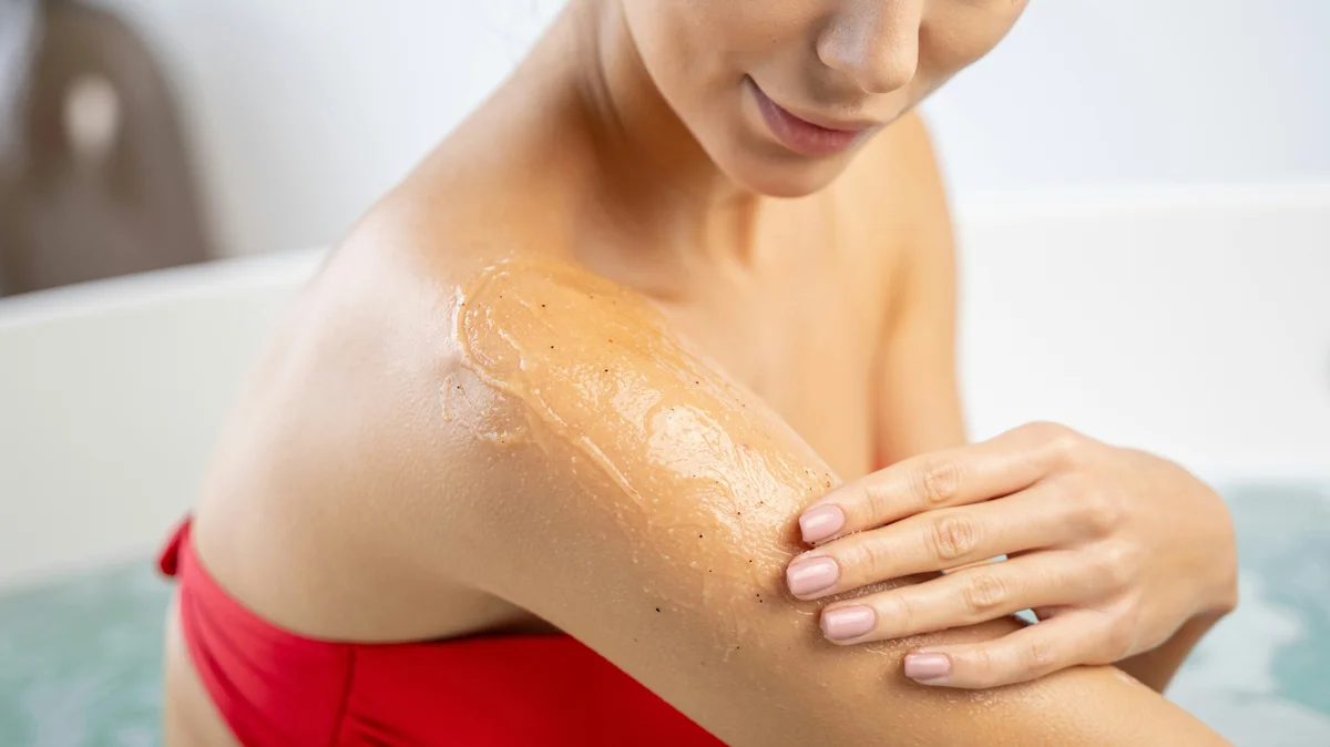 Nahaufnahme einer Frau in einem roten Bikini, die in einem sauberen, weißen Bad ein Peelinggel auf ihren Oberarm aufträgt