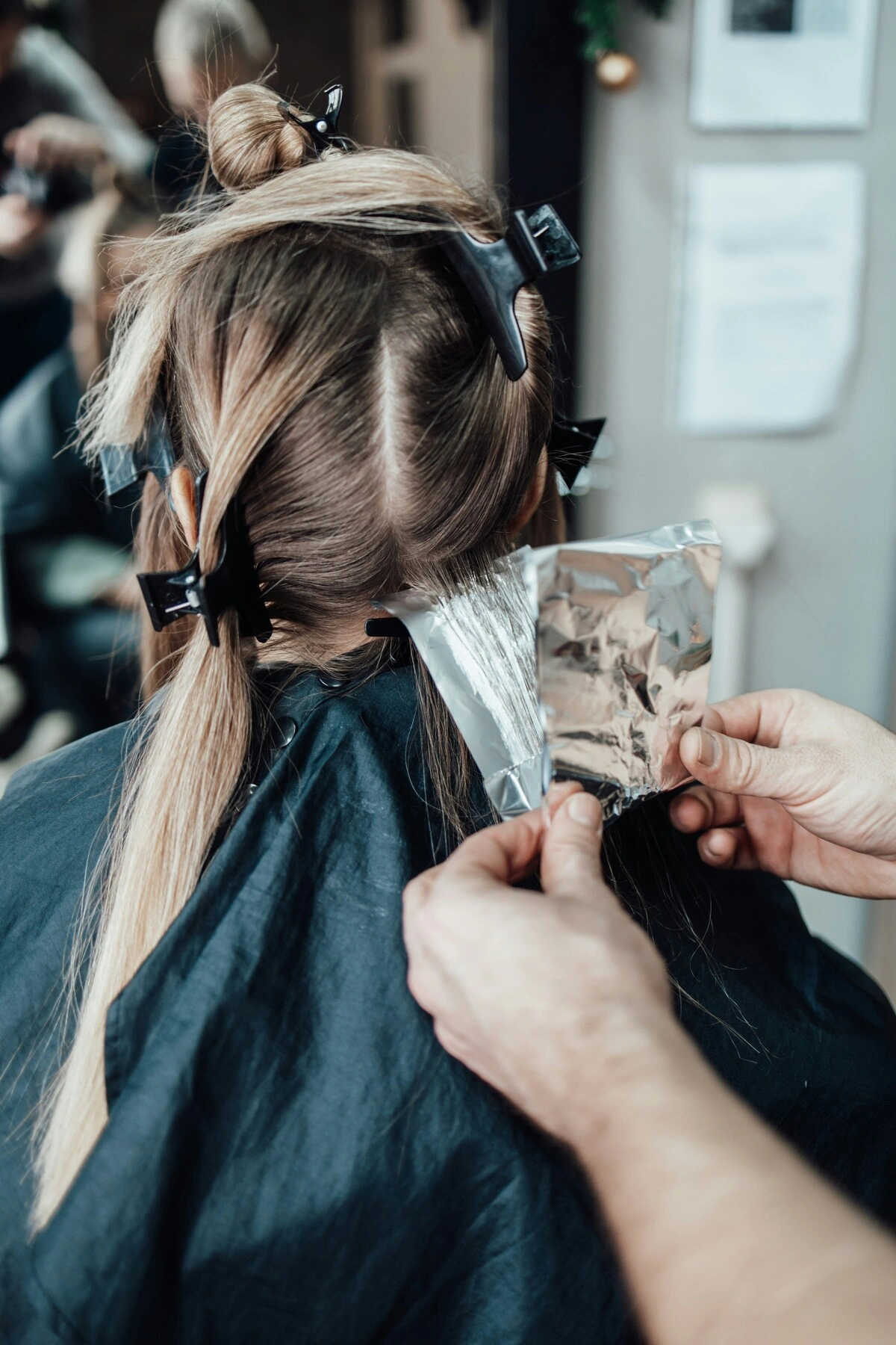 Rückansicht einer Kundin, die auf einem Stuhl sitzt, während eine Friseurin eine Folie in ihr Haar legt, um Strähnchen aufzutragen