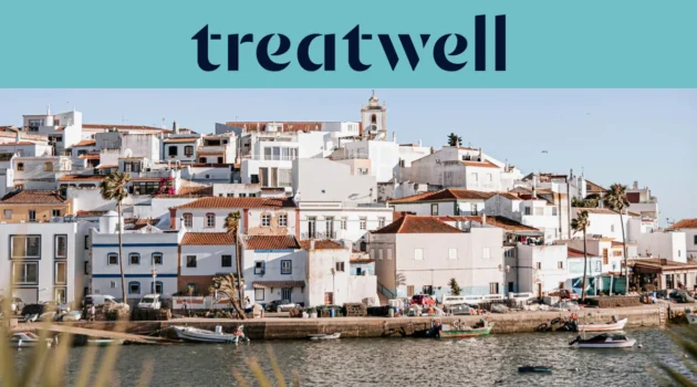 Ein weiß getünchtes Fischerdorf an der Algarve mit terrakottagedeckten Häusern, die einen Hügel über einem ruhigen Gewässer hinaufsteigen, kleinen Fischerbooten am Kai, Palmen und einem Kirchturm unter einem klaren blauen Himmel.