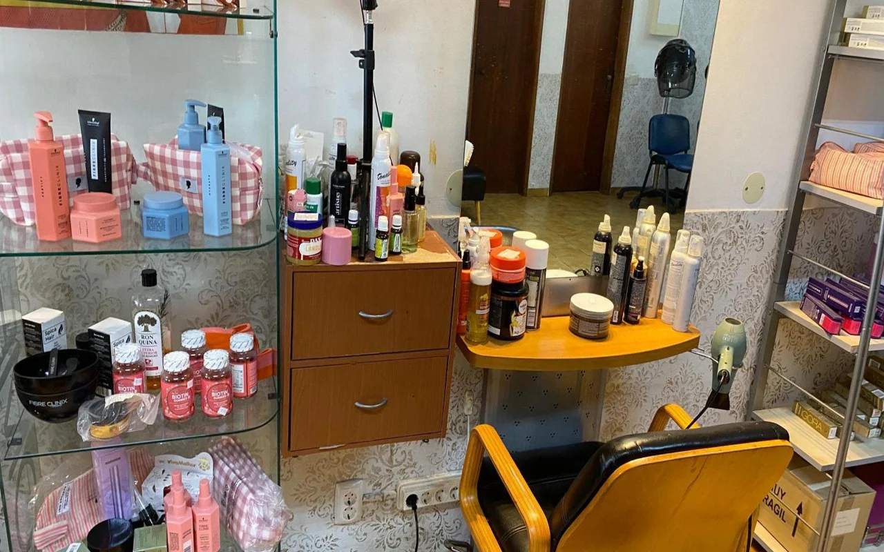 Der Arbeitsplatz einer Friseurin mit einem Wandspiegel und einer Holztheke voller Stylingprodukte, einer Glasvitrine mit Haarpflegeprodukten und Nahrungsergänzungsmitteln daneben und einem klassischen Frisierstuhl in Holz und Schwarz.