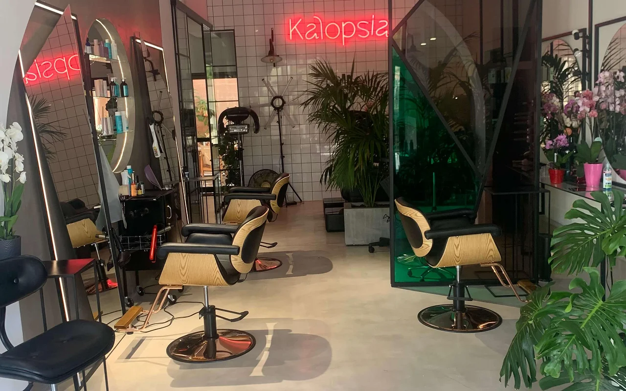 Der Styling-Bereich des Salons Kalopsia in Ermoupoli auf Syros, mit Stühlen in hellem Holz und Schwarz, gewölbten hinterleuchteten Spiegeln, blättrigen Pflanzen und einer rosafarbenen Neonschrift mit dem Wort Kalopsia.