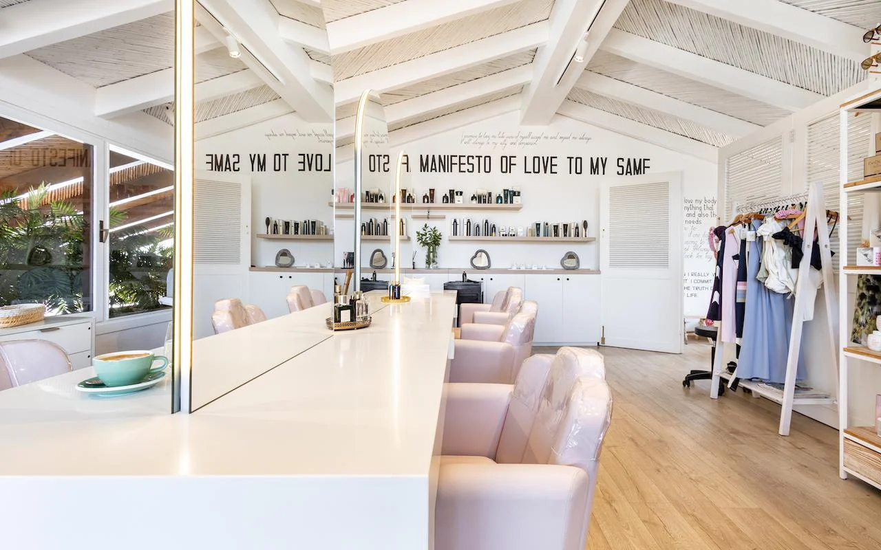 Das helle Interieur von Milk & Roses in Puente Romano, Marbella, mit einem weißen Styling-Tresen, blassrosa Stühlen, einer Balkendecke und Produktregalen entlang der Wand.