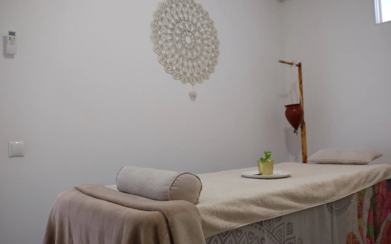 Ein ruhiger Massageraum in sanften, neutralen Tönen, mit einer mit einem Handtuch bedeckten Massageliege und einer Nackenrolle, einem großen gehäkelten Mandala an der Wand und einem Holzständer, der einen hängenden Terrakottatopf hält.
