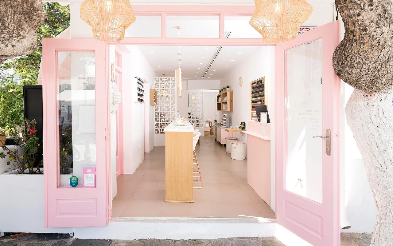 Der rosa gerahmte Eingang von Vivian Nail Boutique in Naoussa auf Paros, mit offenen Türen, geflochtenen Hängeleuchten und einem Holztresen, in einem weiß getünchten kykladischen Gebäude neben einem Baum.
