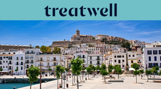 Sonniger Blick über Ibiza-Stadt, mit weiß getünchten Gebäuden, die zur Kathedrale und den alten Festungsmauern von Dalt Vila auf dem Hügel hinaufsteigen. Eine von Bäumen gesäumte Hafenpromenade zieht sich unter einem klaren blauen Himmel durch den Vordergrund.