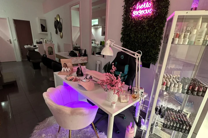 Rosa beleuchteter Beauty-Arbeitsplatz mit einem 'hello gorgeous'-Neonschild auf einer Grünwand, einem cremefarbenen Muschel-Sessel, einem weißen Tisch mit Pinseln und Kirschblüten, und einer Glasvitrine voller bunter Nagellacke.