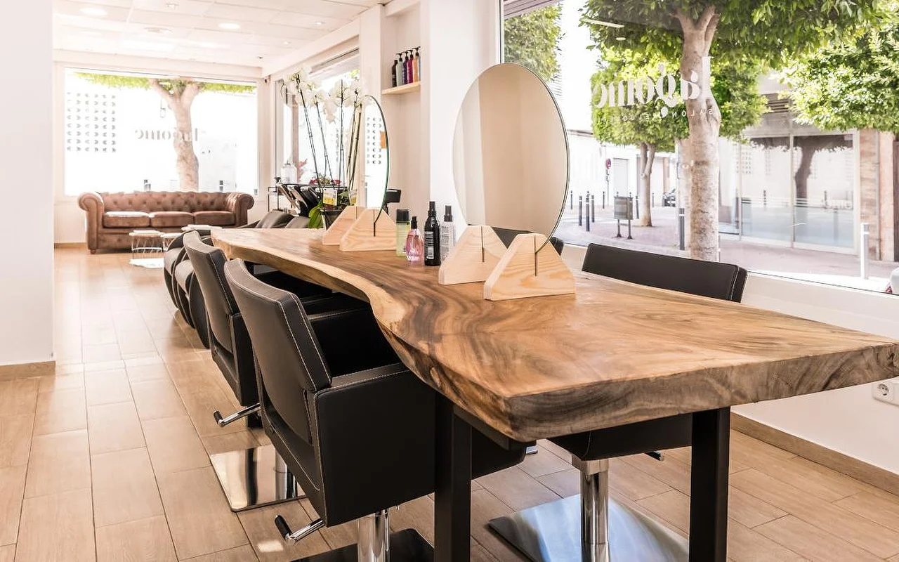 Ein heller Haarsalon, aufgebaut um einen langen Holztresen mit Baumkante und schwarzen Stylingstühlen. Ein runder Spiegel und hölzerne Produktständer stehen auf dem Tresen, ein braunes Chesterfield-Sofa aus Leder wartet im Hintergrund, und hohe Fenster blicken auf eine begrünte Straße.