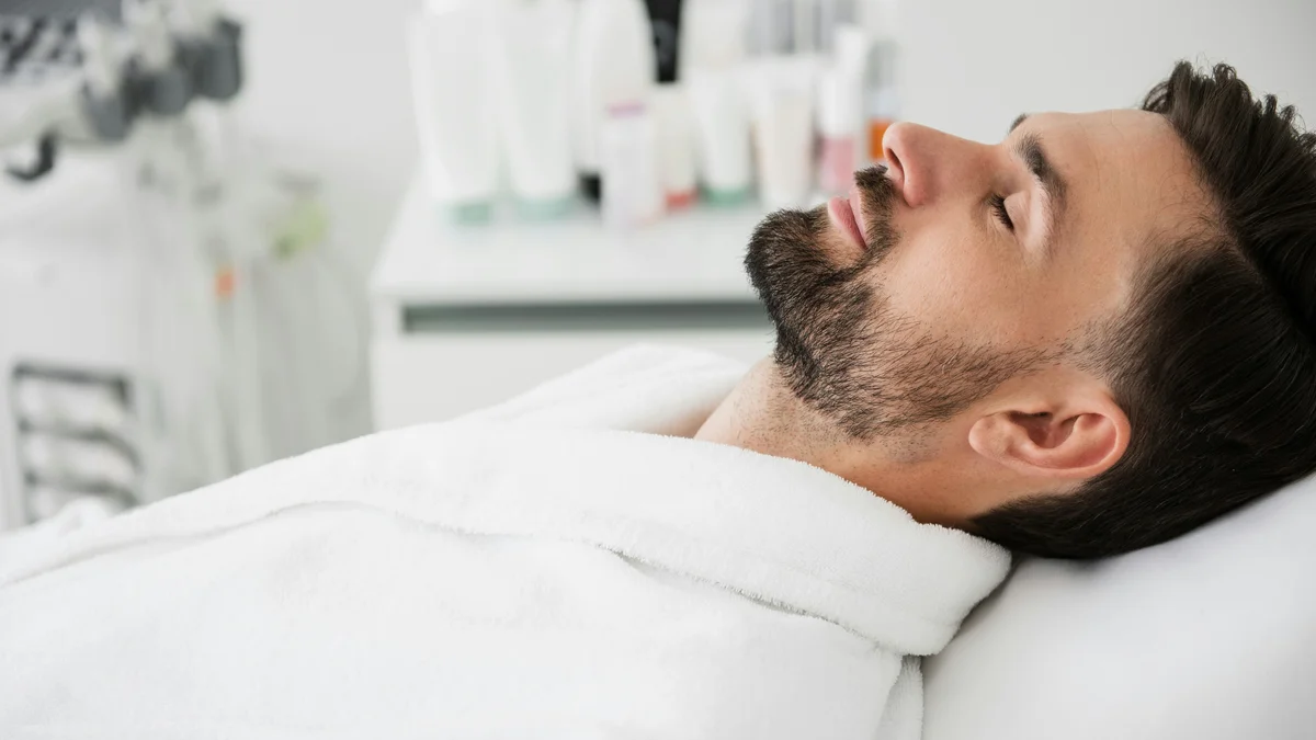 Ein Mann liegt in einem weißen Bademantel auf einer Behandlungsliege, den Kopf nach hinten gelegt und die Augen geschlossen