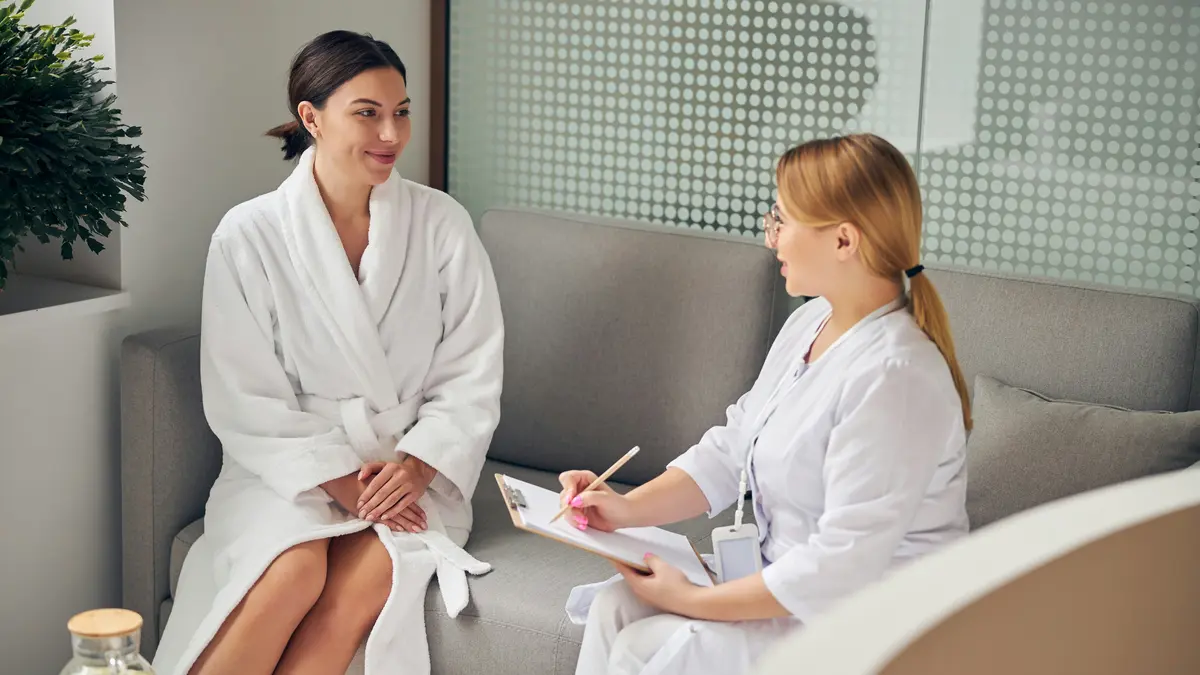 Eine Kundin im weißen Bademantel und eine Fachkraft mit Klemmbrett sitzen nebeneinander auf einem Sofa im Beautysalon, während die Fachkraft Fragen vor der Behandlung stellt und notiert